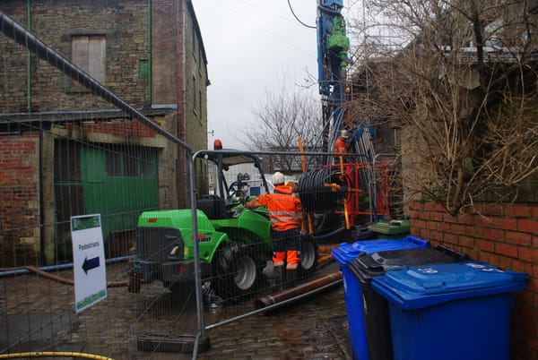 Workers installing a ground loop into a borehole for a ground source heat pump, with machinery in a fenced-off area.